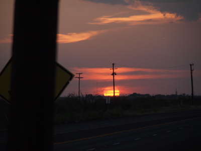 [The sun is a large yellow orb with two thirds of it above the horizon as it sits just behind a power pole. There are some thin pink clouds across the face of it and slightly thicker pink clouds above it. The sky is a purplish blue with some cream colored clouds above it. In the foreground is a small, close-up portion of a power pole with a yellow diamond road sign peeping out on either side.]